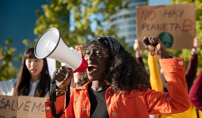 Mulher negra, usando blusa laranja, com megafone grita durante protesto de racismo ambiental.