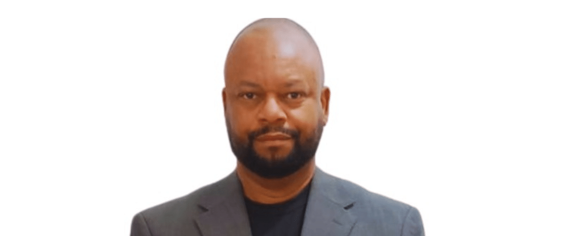 Fundo branco. A imagem mostra o jornalista Welligton, ele é um homem negro, careca com barba e usa termo cinza e camisa preta