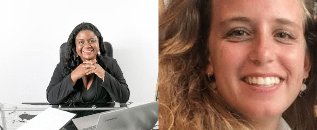 Na esquerda mulher negra, cabelo liso e de roupa social toda preta sorri sentada em uma mesa. À direita, mulher de cabelo encaracolado, loira, branca sorri.