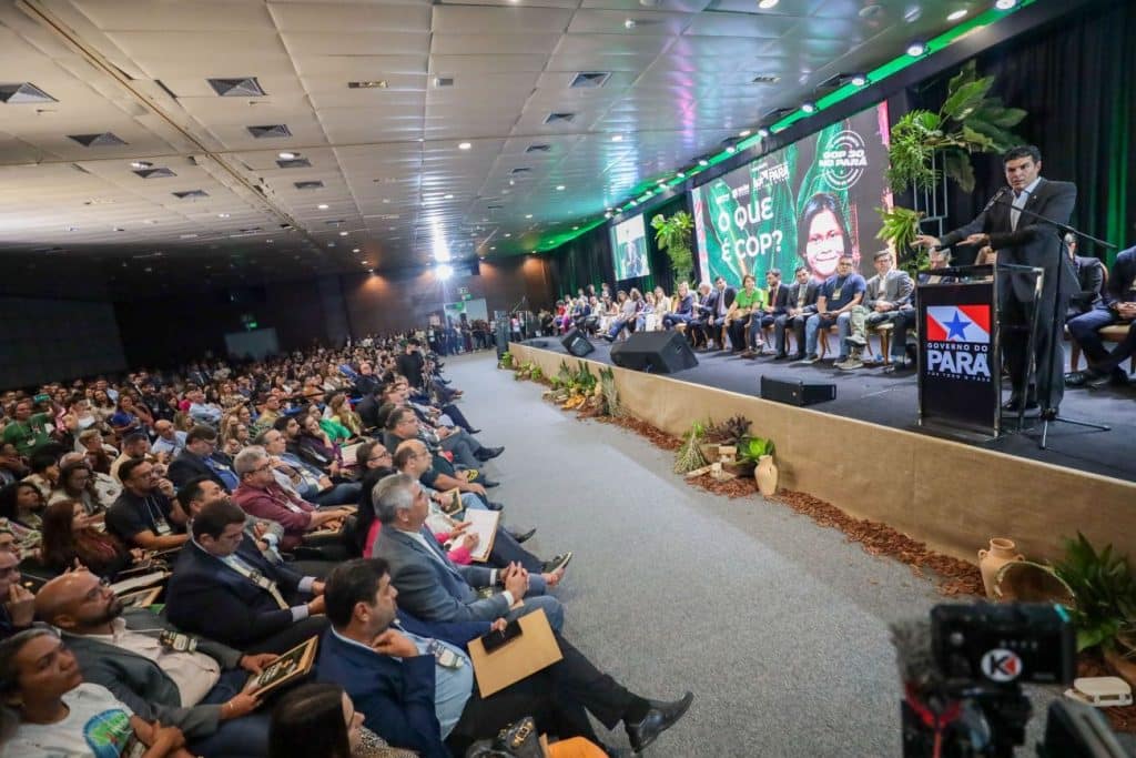 Foto mostra um homem engravatado, indígena, mas de terno, discursando em um púlpito em um palco no canto esquerdo. Atrás está um telão verde com o nome COP30, a visibilidade de um indígena e paisagens ambientais, e do lado esquerdo da foto está uma fila de participantes sentados em cadeiras (aproximadamente 25 a 40), o escutando, no palco, todos de roupas sociais e formais. 

Na parte inferior da foto, aparece uma plateia cheia, em um auditório.